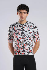 A man wearing a short-sleeve cycling jersey with a black, white, and red pixelated camouflage pattern, paired with black cycling shorts, against a light gray studio background.