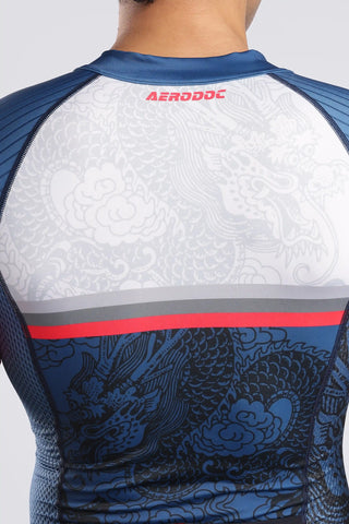 A close-up back view of a man wearing a short-sleeve tyrisuit with a white upper section, blue lower section, and dragon pattern. The aerodoc trisuit features a horizontally red 'AERODOC' logo on the back