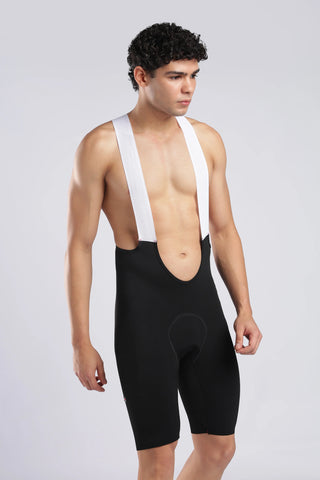 A male model wearing black cycling bib shorts with white shoulder straps, photographed against a light gray studio background.