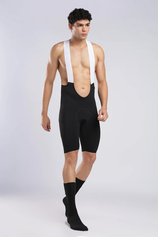 A male model wearing black cycling bib shorts with white shoulder straps, paired with black crew socks, standing against a light gray studio background.