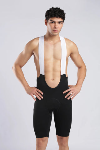 A man modeling black compression shorts with white suspenders against a light gray studio background.
