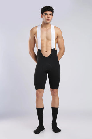 A male model wearing black cycling shorts with white shoulder straps, paired with black crew socks, standing against a light gray studio background.
