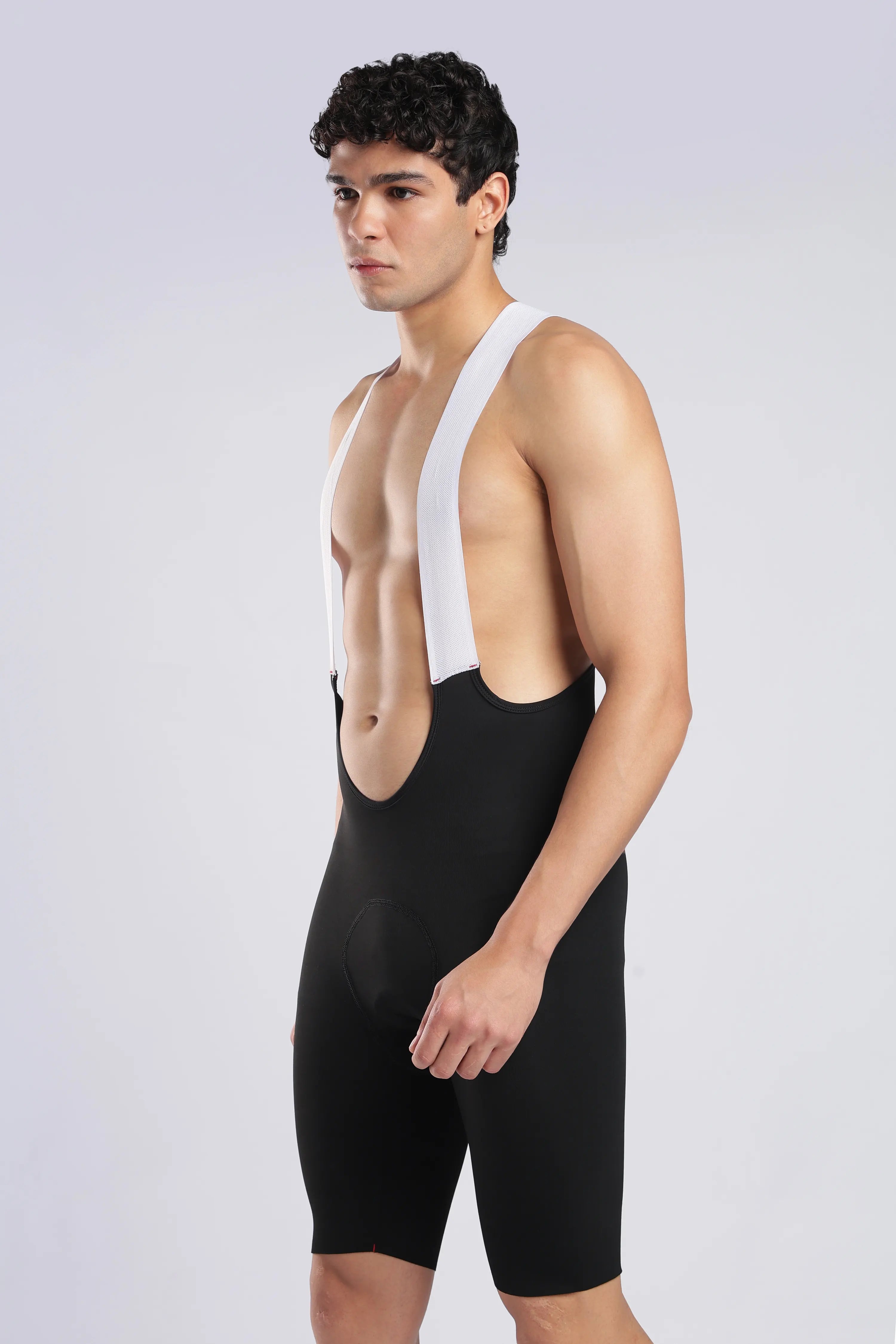 A man modeling a black bib shorts and suspenders set against a light gray studio background.