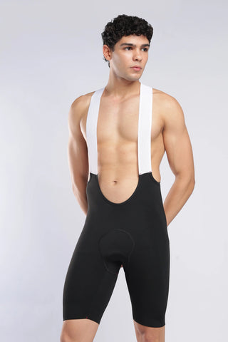 A male model wearing black cycling bib shorts with wide white shoulder straps, photographed against a light gray studio background.