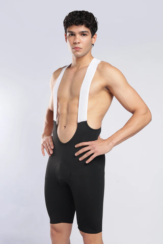 A male model wearing a black athletic bib shorts and white shoulder straps, standing against a light gray studio background.