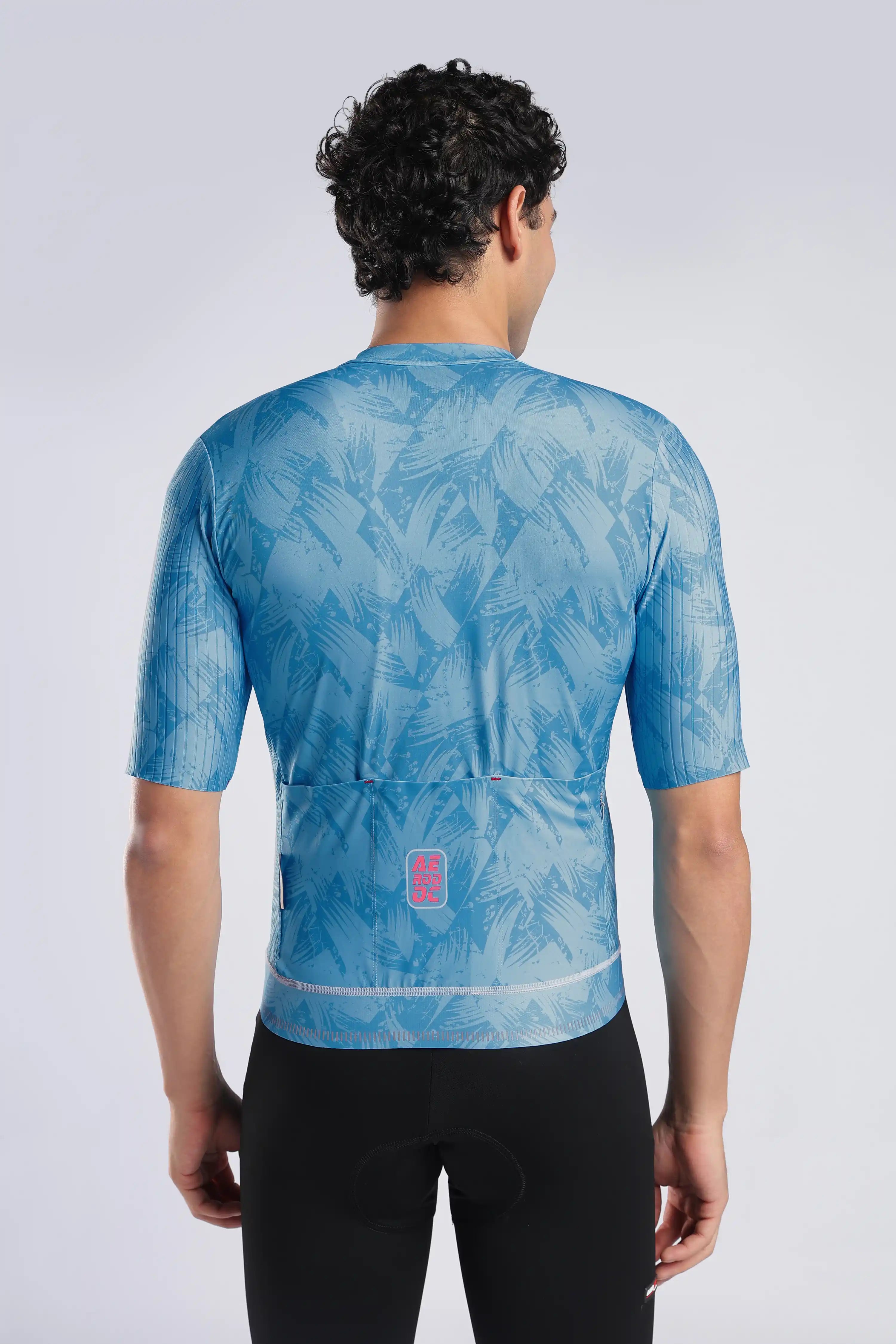 A man viewed from behind wearing a light blue short-sleeve cycling jersey with a tropical leaf pattern and black cycling shorts against a light gray studio background.