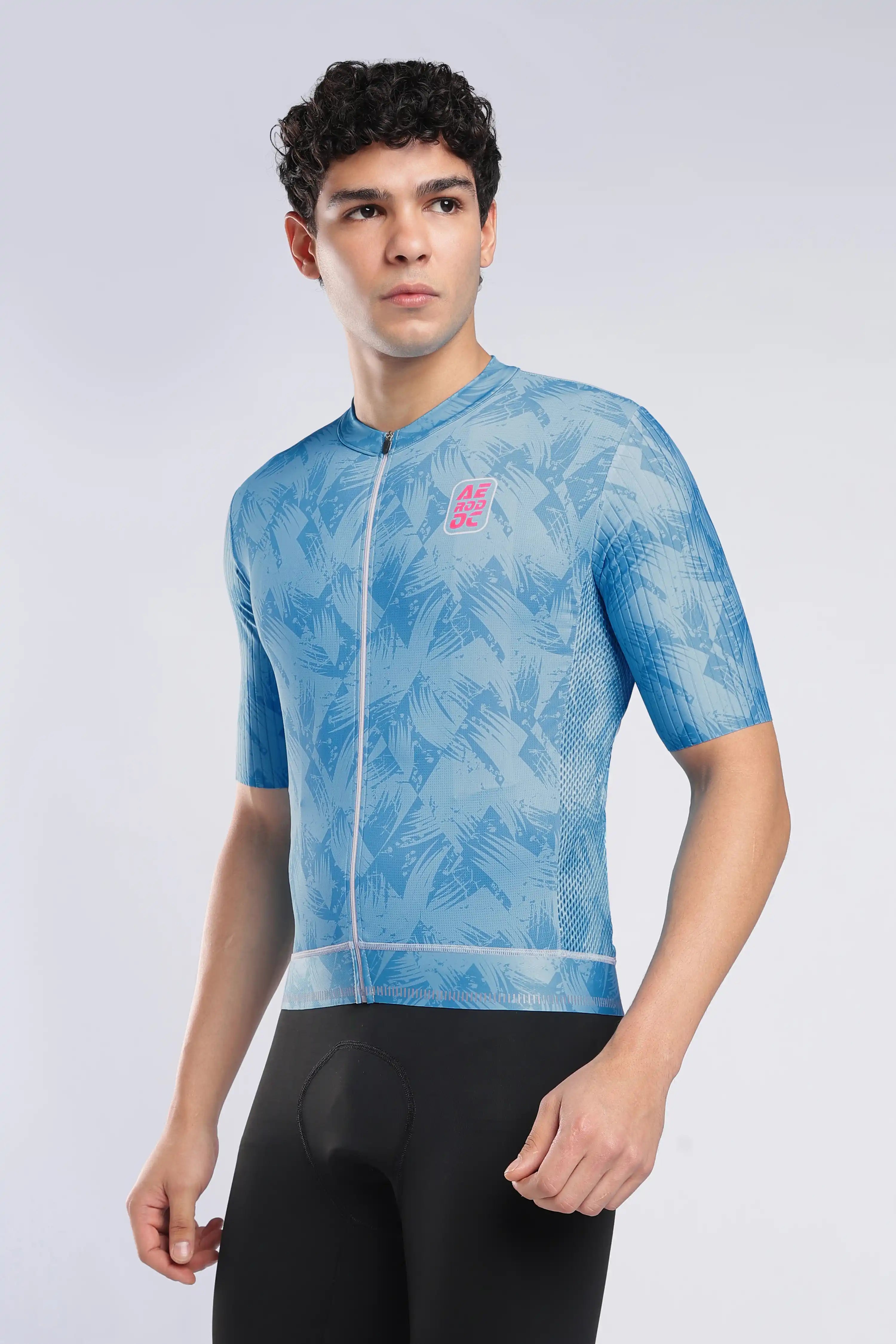 A man wearing a light blue short-sleeve cycling jersey with a subtle palm leaf pattern and a pink logo on the left chest, paired with black cycling shorts, against a light gray studio background.