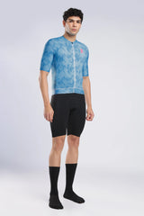 A male model wearing a light blue patterned short-sleeve cycling jersey, black cycling shorts, and black crew socks, standing against a light gray studio background.