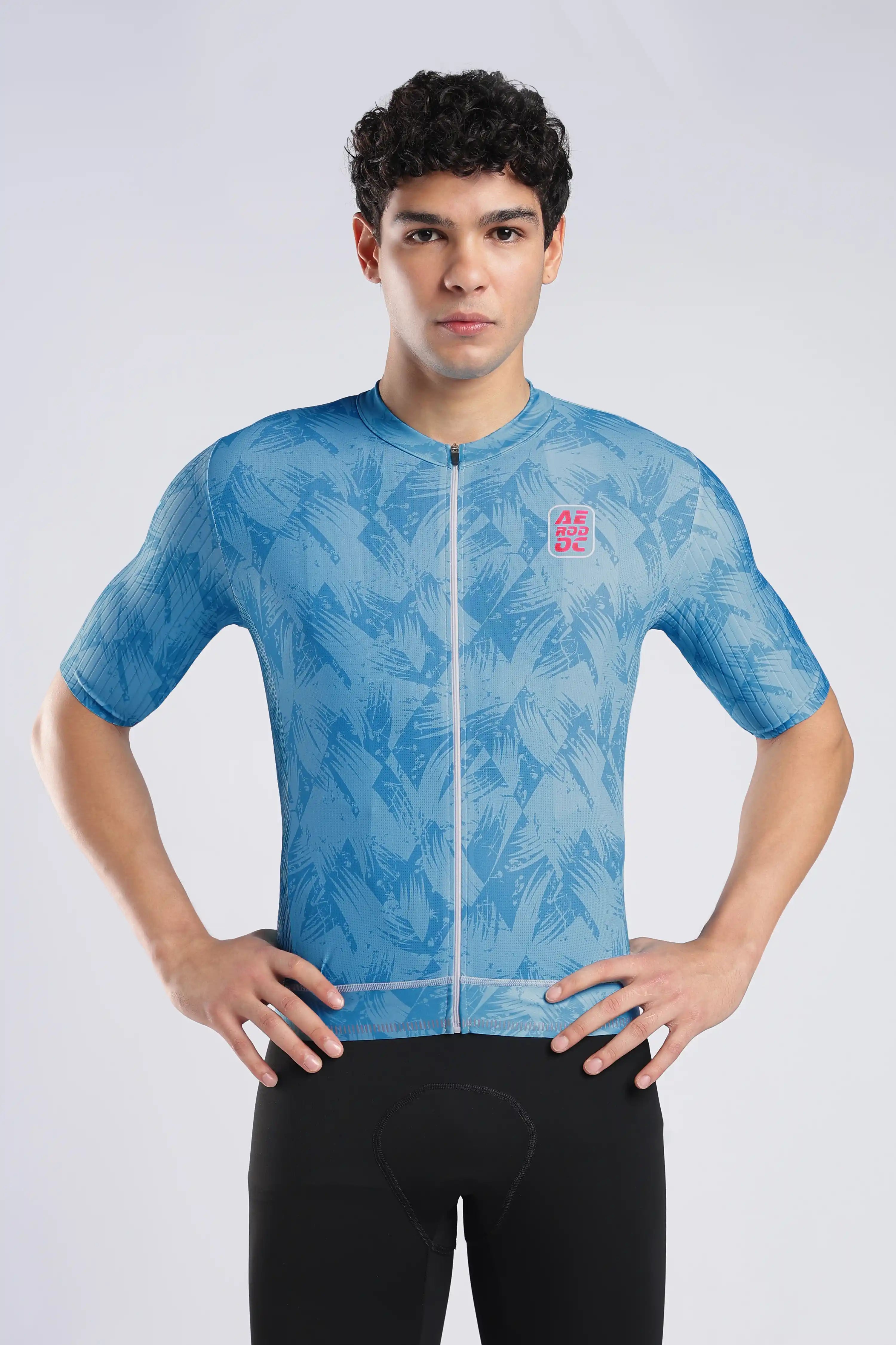 A man wearing a light blue short-sleeve cycling jersey with a subtle tropical leaf pattern and a small red logo on the left chest, paired with black cycling shorts, against a light gray studio background.
