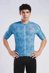 A man wearing a light blue short-sleeve cycling jersey with a subtle tropical leaf pattern and a small red logo on the left chest, paired with black cycling shorts, against a light gray studio background.