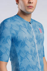 A close-up of a man wearing a light blue short-sleeve cycling jersey with a darker blue abstract leaf pattern and a small red and white AERODOC logo on the chest.