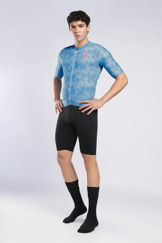 A male model wearing a light blue patterned short-sleeve cycling jersey, black cycling shorts, and black crew socks, standing against a light gray studio background.