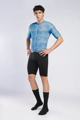 A male model wearing a light blue patterned short-sleeve cycling jersey, black cycling shorts, and black crew socks, standing against a light gray studio background.