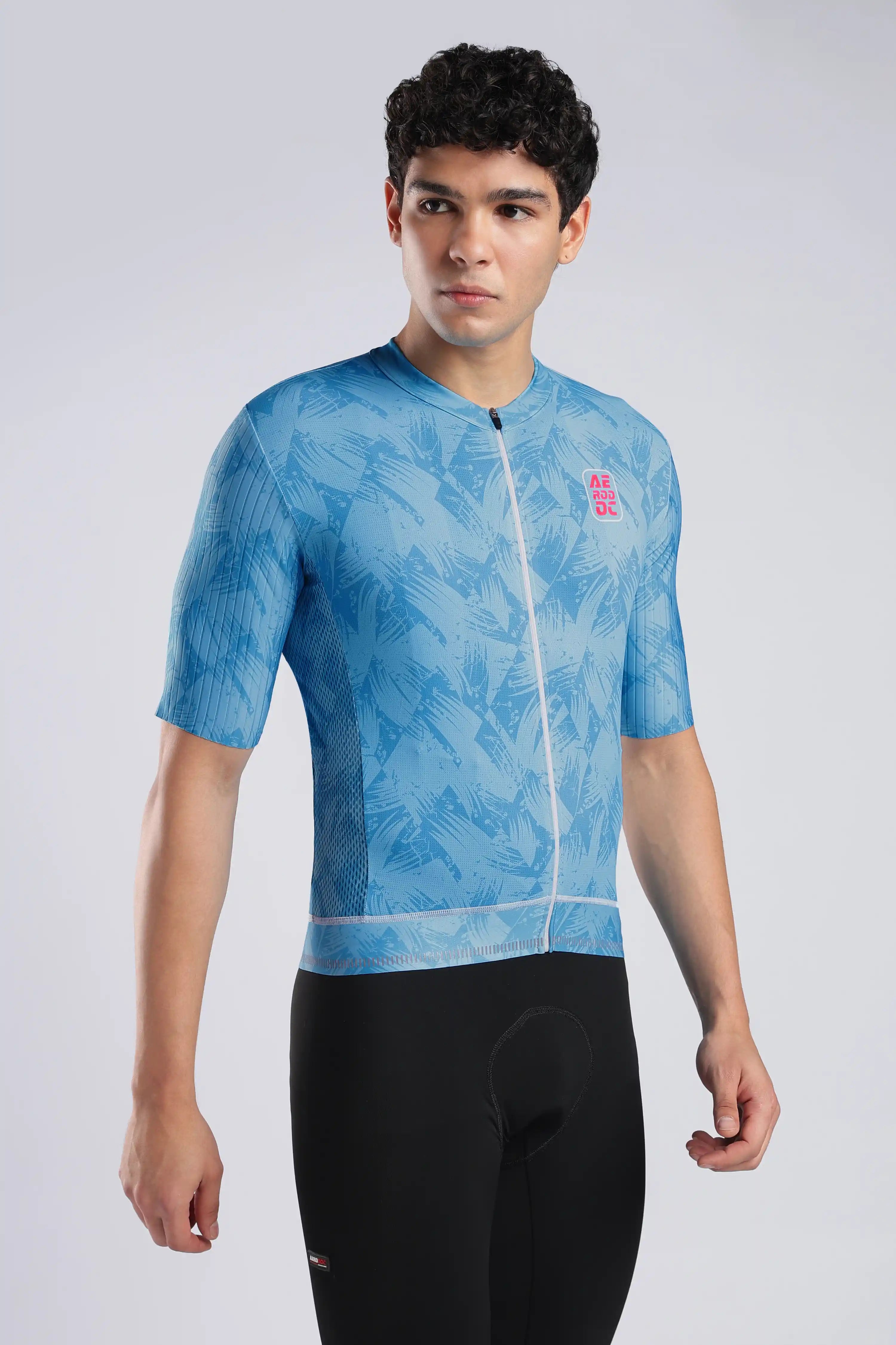 A man modeling a light blue short-sleeve cycling jersey with a subtle leaf pattern and a small pink logo on the chest, paired with black cycling shorts against a light gray studio background.