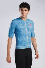 A man modeling a light blue short-sleeve cycling jersey with a subtle leaf pattern and a small pink logo on the chest, paired with black cycling shorts against a light gray studio background.