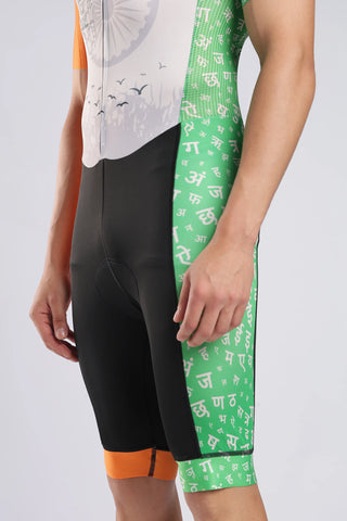 Close-up of a person wearing AERODOC trisuit with a color-blocked design: black on the left leg, green on the right leg with white Devanagari script, and orange at the lower hem, paired with a white and orange cycling jersey against a light gray background.