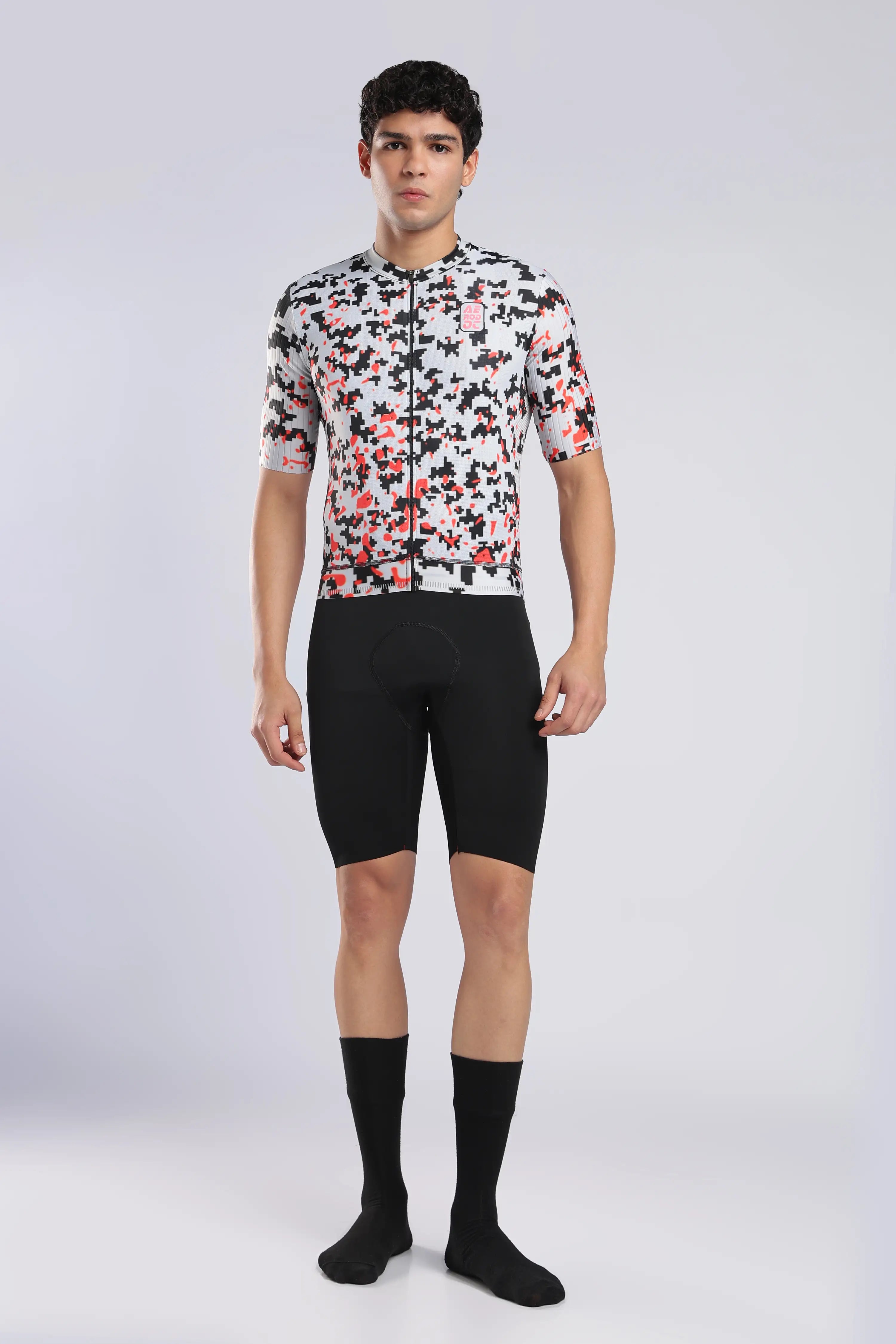 Aerodoc glitch cycling jersey a male model wearing a short-sleeve cycling jersey with a white, black, and red abstract pattern, paired with black cycling shorts, black socks, and black shoes, standing against a light gray studio background.