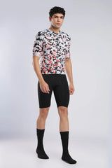 A male model wearing a short-sleeve cycling jersey with a black, white, and red abstract pattern, paired with black cycling shorts and black crew socks, standing against a light gray studio background.