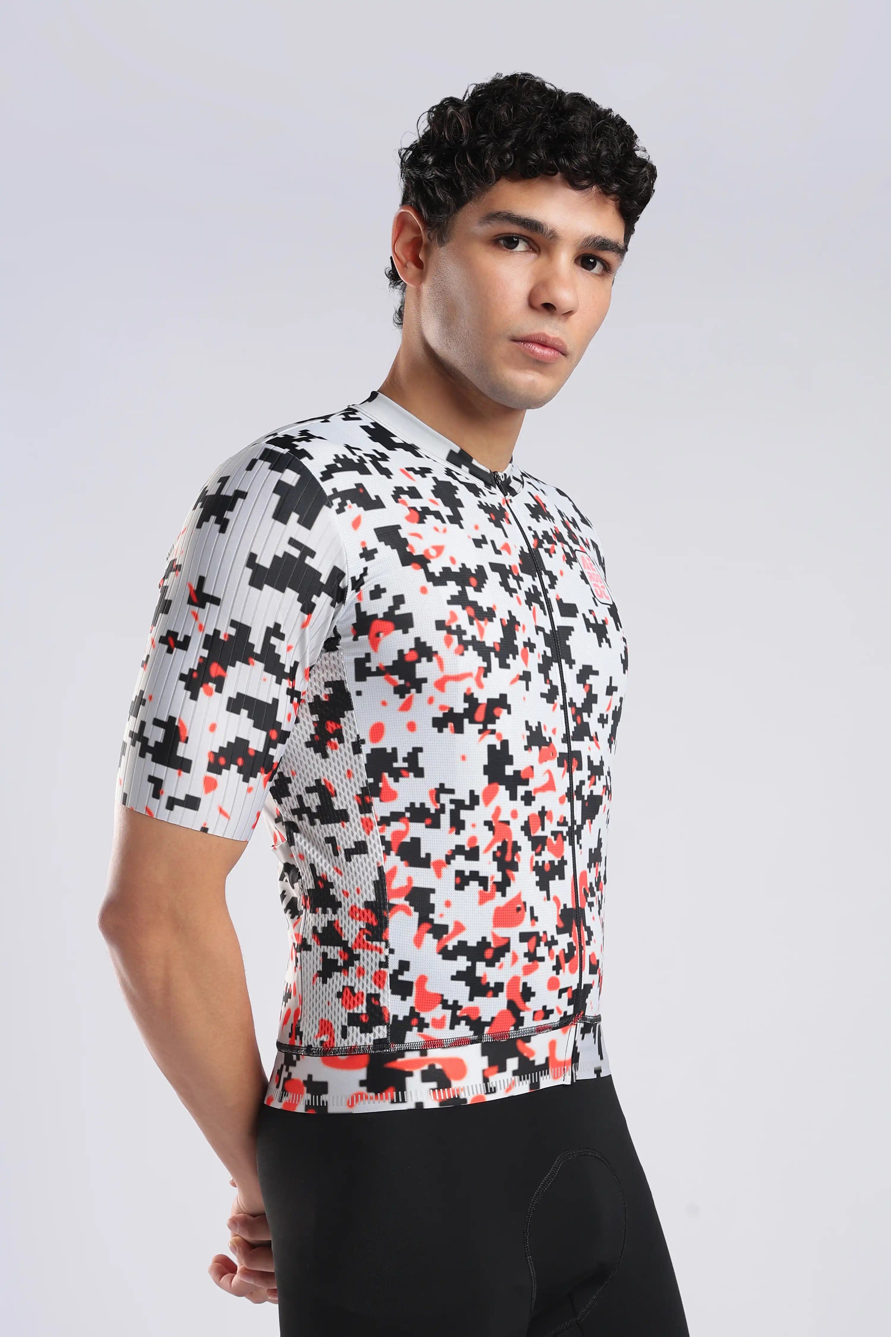 A man wearing a short-sleeve aerodoc glitch cycling jersey with a black, white, and red pixelated pattern, paired with black cycling shorts, photographed against a light gray studio background.