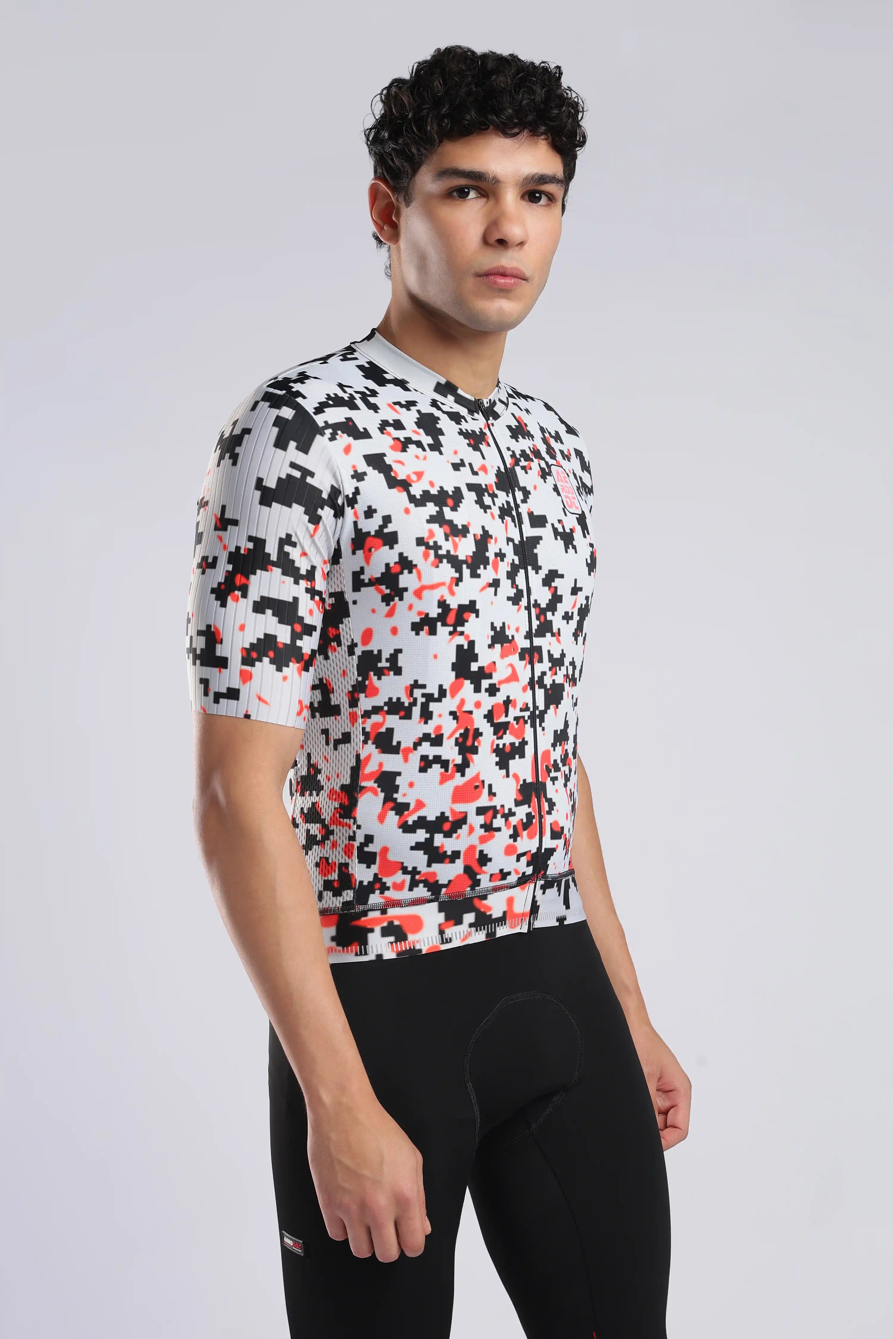A man wearing a short-sleeve aerodoc glitch cycling jersey with a black, white, and red pixelated camouflage pattern, paired with black cycling shorts, against a light gray studio background.