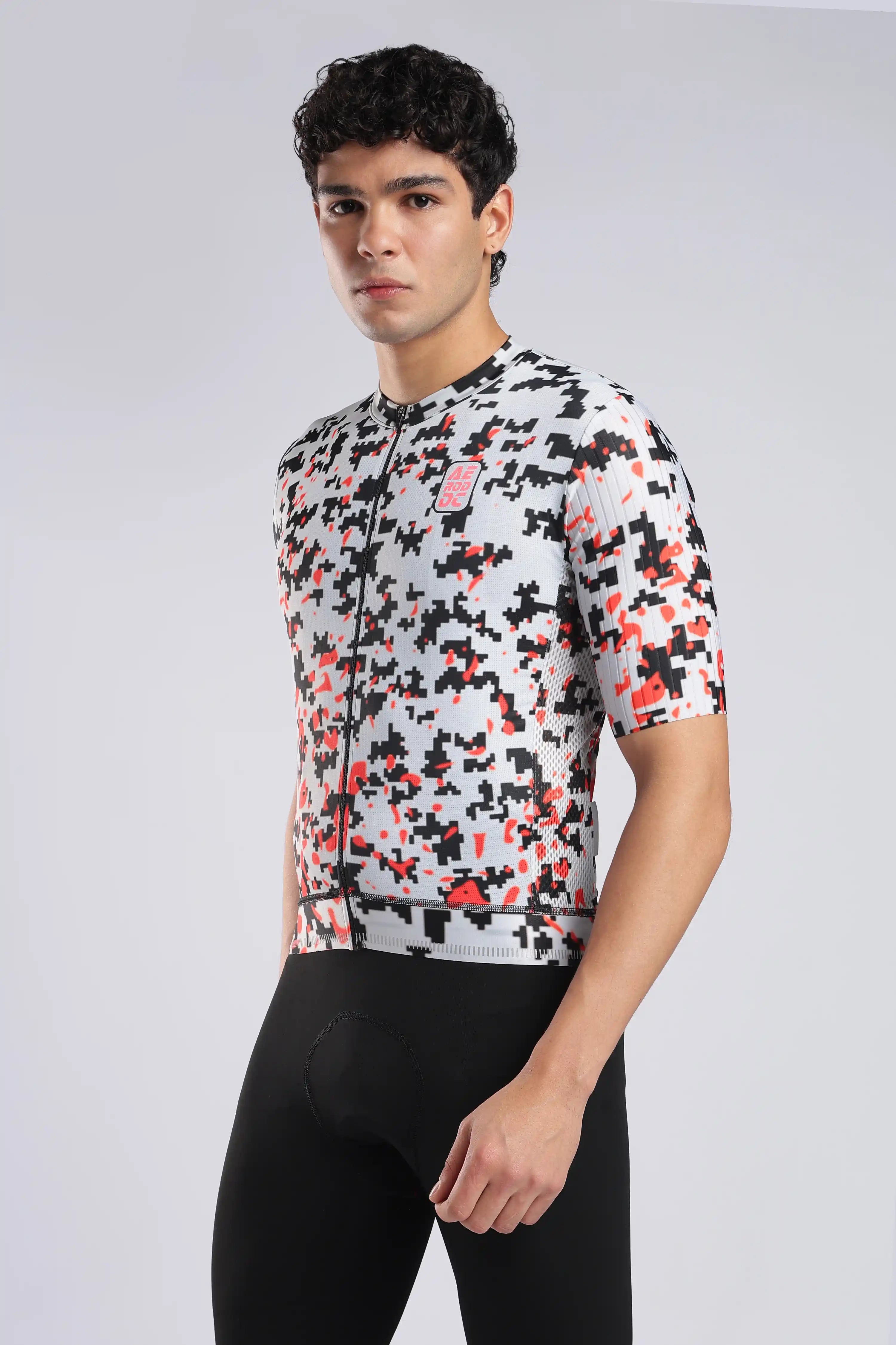A man wearing a short-sleeve aerodoc glitch cycling jersey with a black, white, and red geometric pixelated pattern, paired with black cycling shorts, against a light gray studio background.