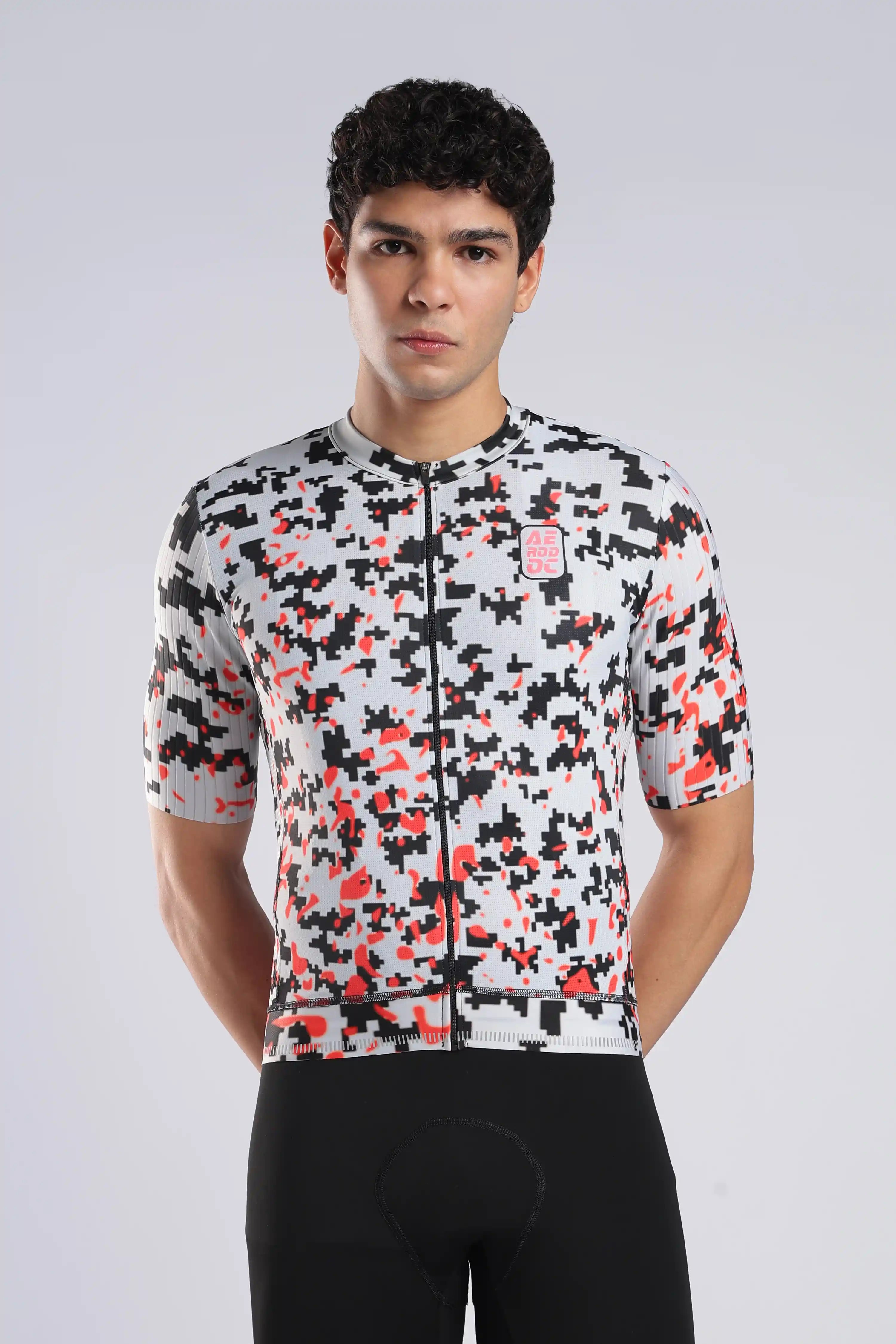 A man wearing a short-sleeve cycling jersey with a black, white, and red pixelated camouflage pattern, paired with black cycling shorts, against a light gray studio background.