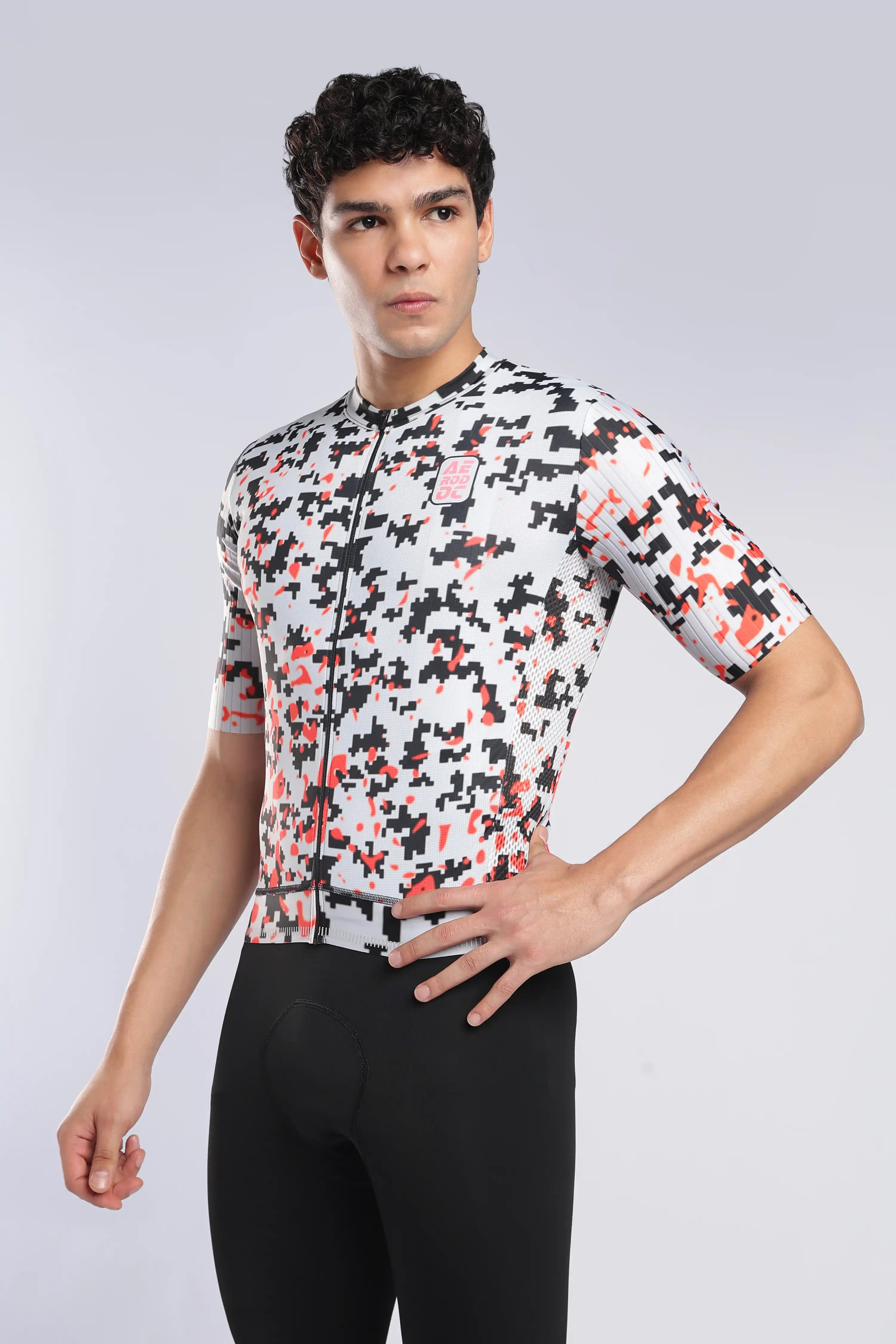 A man modeling a short-sleeve cycling jersey with a white, black, and red camouflage pattern, paired with black cycling tights, against a light gray studio background.