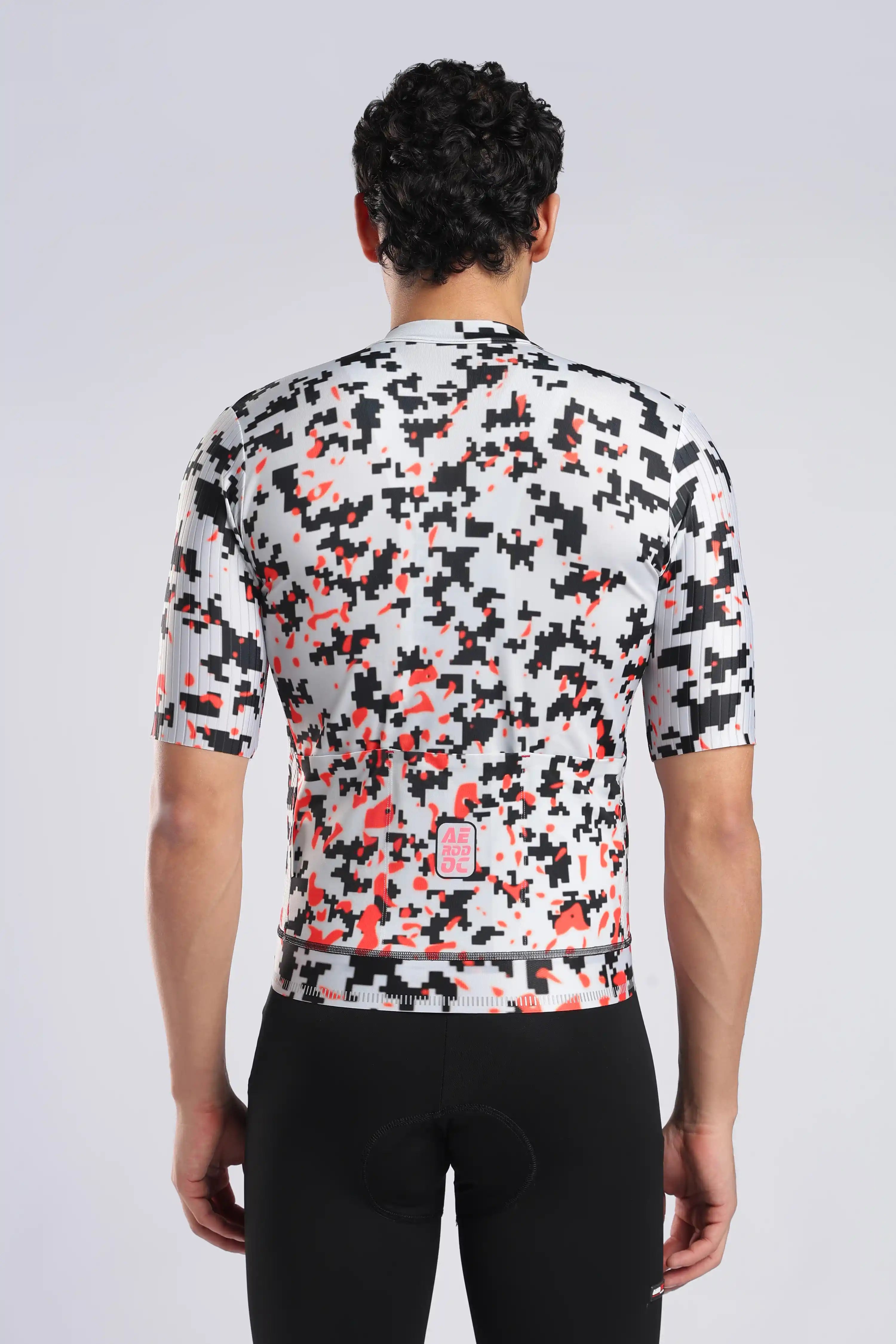 Rear view of a man wearing a short-sleeve aerodoc cycling jersey with a black, white, and orange abstract pixelated pattern, paired with black cycling shorts against a light gray studio background.