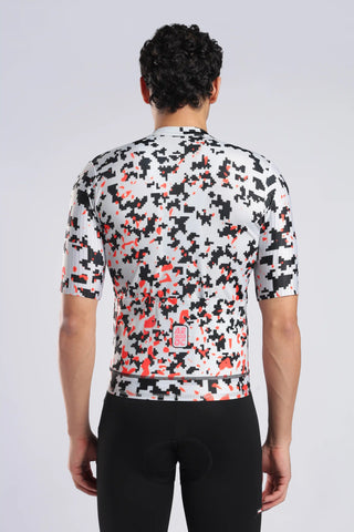 Rear view of a man wearing a short-sleeve aerodoc cycling jersey with a black, white, and orange abstract pixelated pattern, paired with black cycling shorts against a light gray studio background.