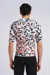Rear view of a man wearing a short-sleeve aerodoc cycling jersey with a black, white, and orange abstract pixelated pattern, paired with black cycling shorts against a light gray studio background.
